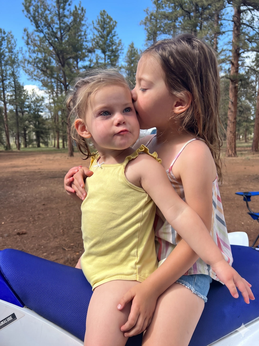 Two young Bear Basics daughters outdoors, the older sister kissing her little sister on the cheek